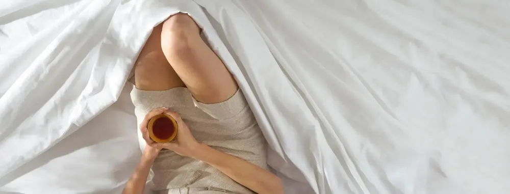 Overhead view of the legs of a person sitting in a pile of sheets and holding a cup of coffee